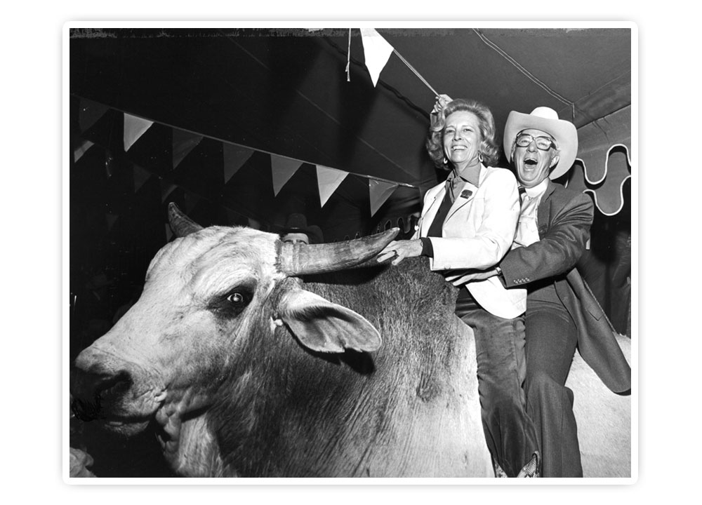 Mr. John S. Justin, Jr. and Mrs. Jane Chilton Justin pose on a Bucking Bull, smiling and laughing, holding each other.